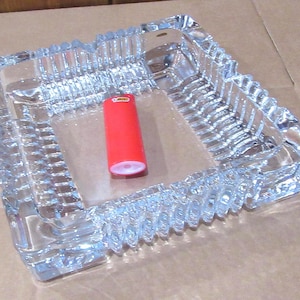 Könnte beinhalten: Ein klarer, quadratischer Glasaschenbecher mit strukturiertem, geriffeltem Design. Ein rotes Feuerzeug liegt im Aschenbecher. Der Aschenbecher besteht aus dickem Glas und hat ein dekoratives, geometrisches Muster.