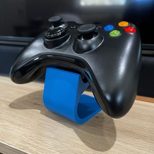 May include: A black video game controller resting on a bright blue stand. The controller has colored buttons and two joysticks. The stand is curved and made of a solid material. The setup is on a light-colored wooden surface.