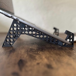 May include: Black tablet stand with a honeycomb design. The stand is angled to hold a tablet and has a small lip to keep the tablet in place. The stand is made of a matte black material.
