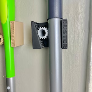 Puede incluir: Dos herramientas de limpieza están montadas en una pared con un soporte de plástico negro. La herramienta de la izquierda es verde y está etiquetada como "SWIFFER". La herramienta de la derecha es gris y está etiquetada como "DUSTER".