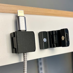 May include: A black cable management clip with a magnetic closure. The clip is attached to a white surface and is holding a white and gray braided cable with a silver lightning connector.