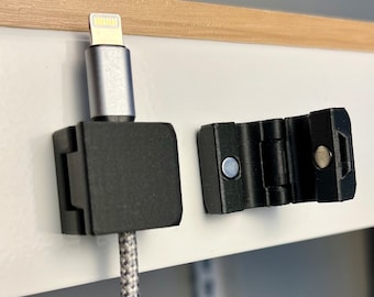 Könnte beinhalten: Ein schwarzer Kabelmanagementclip mit magnetischem Verschluss. Der Clip ist an einer weißen Oberfläche befestigt und hält ein weiß-graues geflochtenes Kabel mit einem silbernen Lightning-Stecker.