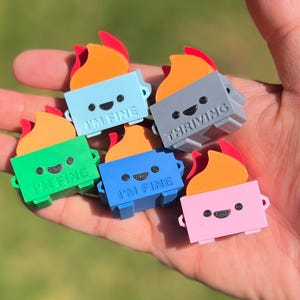 May include: A collection of colorful, miniature campfire figurines. Each figurine features a rectangular base with a smiling face and a flame design. The bases are in various colors, including blue, green, gray, pink, and blue. The text "I'M FINE" or "THRIVING" is printed on the base.