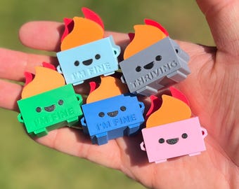 May include: A collection of colorful, miniature campfire figurines. Each figurine features a rectangular base with a smiling face and a flame design. The bases are in various colors, including blue, green, gray, pink, and blue. The text "I'M FINE" or "THRIVING" is printed on the base.