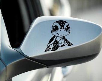 以下が含まれることがあります： 車のサイドミラーに貼られた、漫画のウミガメの黒いビニールデカール。カメは大きな目、笑顔の口、模様のある甲羅をしています。このデカールは、車両用の装飾アクセサリーです。
