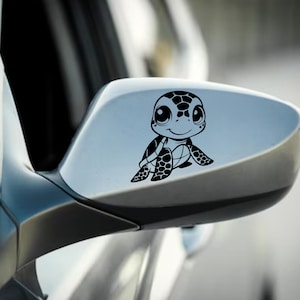 May include: A black vinyl decal of a cartoon sea turtle is affixed to a car's side mirror. The turtle has large eyes and a smiling expression. The decal is a decorative accessory for vehicles.