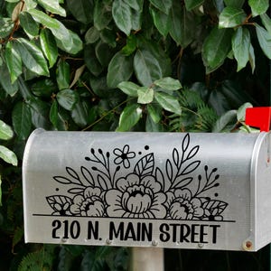 May include: A silver mailbox with a black floral design and the address "210 N. MAIN STREET". The mailbox has a red flag and is set against a backdrop of green foliage.