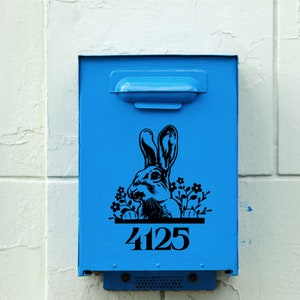 Op de afbeelding: Een helderblauwe brievenbus met een zwart konijn- en bloemmotief. Het nummer 4125 staat in het zwart onderaan. De brievenbus is bevestigd aan een wit en lichtblauw muur.