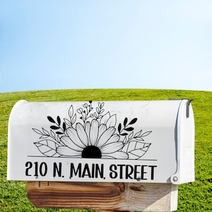 May include: White mailbox with a black floral design and the address "210 N. MAIN STREET" printed on the front. The mailbox is mounted on a wooden post, set against a backdrop of green grass and a blue sky.