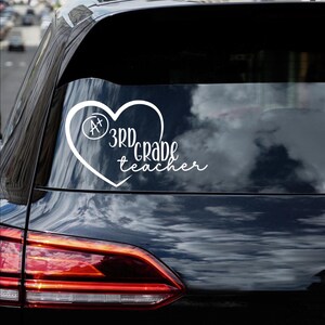May include: White vinyl decal featuring a heart shape with the text "3rd Grade teacher". Inside the heart is a circle with the letters "A+". The decal is designed to be applied to a car window or similar surface.
