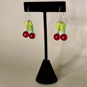 May include: A pair of dangle earrings with red cherry charms and green leaf accents. The earrings are hanging from a black earring stand.