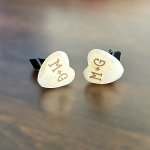 May include: Two white heart-shaped cufflinks with black metal clasps. The cufflinks are engraved with the initials "M+G".