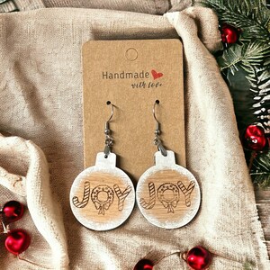 May include: A pair of wooden Christmas ornament earrings with the word "JOY" and a wreath design. The earrings are white with a wood grain texture and have silver hooks. The earrings are on a brown card with the text "Handmade with love".