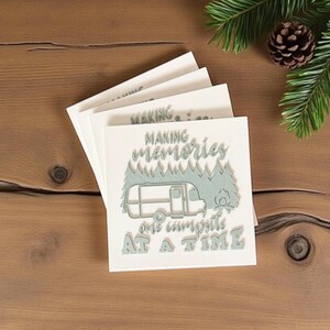 May include: A stack of square, off-white coasters with a camping theme. Each coaster features a light teal design with the words "MAKING MEMORIES ONE CAMPSITE AT A TIME" and an illustration of a camper, trees, and a campfire.