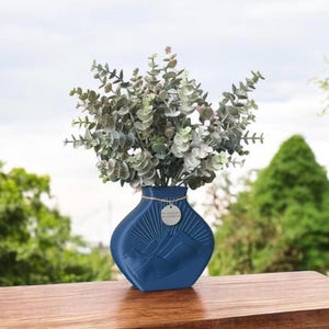 May include: A blue ceramic vase with a mountain and sun design. The vase is filled with a bouquet of green eucalyptus leaves.
