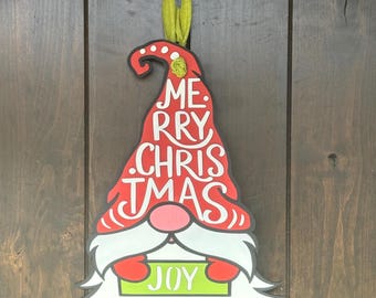 May include: Red and white gnome ornament with a green hat and a sign that says "Joy". The hat has the words "Merry Christmas" written on it.
