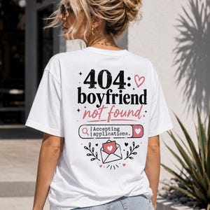 Könnte beinhalten: Weißes T-Shirt mit dem Text "404: boyfriend not found" in Schwarz und Rosa. Das Design beinhaltet eine Suchleiste mit "Accepting applications" und einen Umschlag mit einem Herz. Das T-Shirt wird von einer Person mit blonden Haaren getragen.