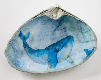 May include: A blue whale illustration inside a white seashell with a pearl on top. The seashell is decorated with a blue and white pattern.