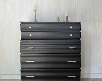 May include: A black dresser with five drawers and gold hardware. The dresser has a modern design with rounded edges and sits on four gold legs.