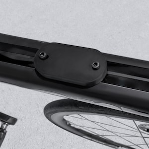 Puede incluir: Primer plano de un cuadro de bicicleta negro con una cubierta ovalada negra asegurada con dos tornillos. La imagen muestra una porción de la rueda y el diseño elegante del cuadro, lo que sugiere componentes y accesorios de ciclismo.