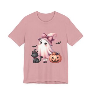 May include: A light pink t-shirt featuring a Halloween-themed illustration. The design includes a cute ghost wearing a pink witch hat with a bow, a black cat, a carved pumpkin, and bats. The artwork is in a watercolor style.