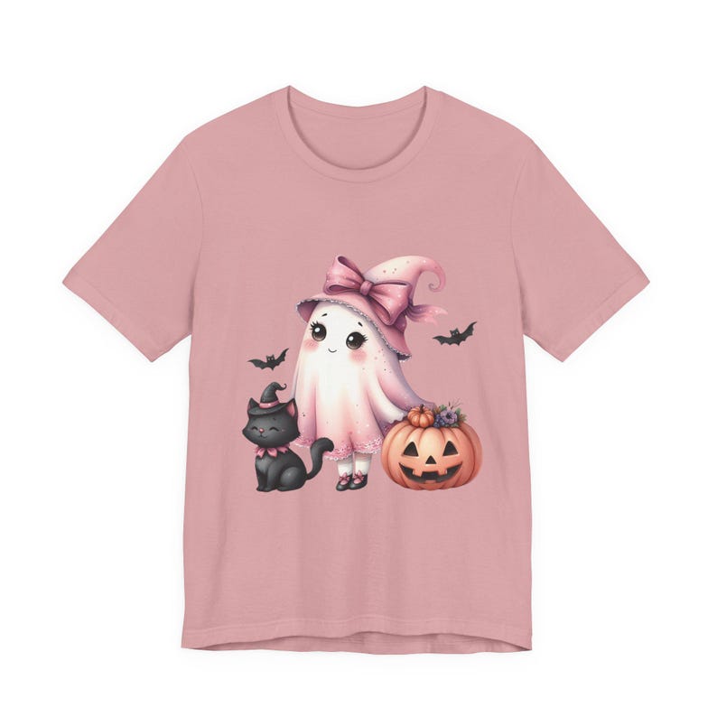 May include: A light pink t-shirt featuring a Halloween-themed illustration. The design includes a cute ghost wearing a pink witch hat with a bow, a black cat, a carved pumpkin, and bats. The artwork is in a watercolor style.