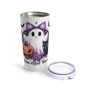 May include: A white travel mug with a Halloween-themed design. The design features a cartoon ghost with a purple bow, a jack-o'-lantern, and a black cat. The words "Boo-tiful Brew" are printed on the mug.