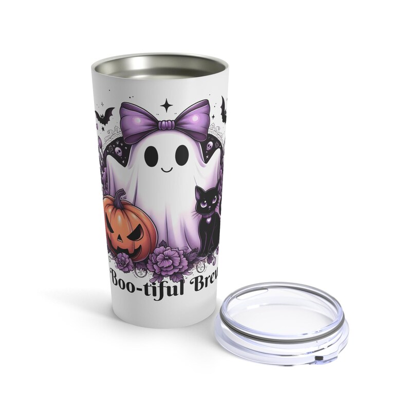 May include: A white travel mug with a Halloween-themed design. The design features a cartoon ghost with a purple bow, a jack-o'-lantern, and a black cat. The words "Boo-tiful Brew" are printed on the mug.