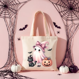 May include: A cream-colored tote bag featuring a Halloween-themed illustration. The design includes a cartoon ghost wearing a pink witch hat and bow, a black cat, and a carved pumpkin. Bats and spiders decorate the background.