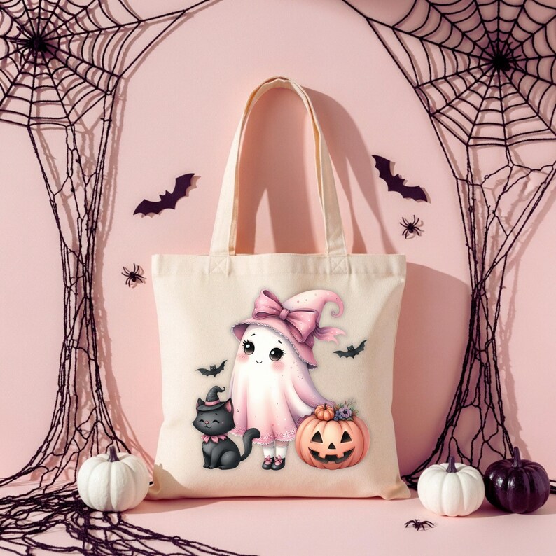 May include: A cream-colored tote bag featuring a Halloween-themed illustration. The design includes a cartoon ghost wearing a pink witch hat and bow, a black cat, and a carved pumpkin. Bats and spiders decorate the background.
