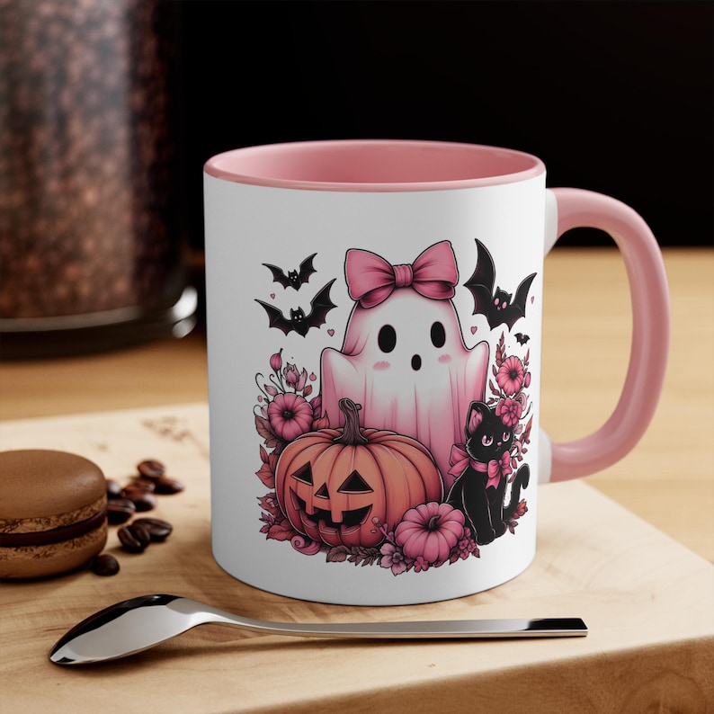 The mug features a pink ghost with a pink bow, a black cat with a pink bow, a pumpkin with a jack-o-lantern face, and pink flowers.