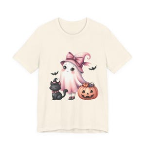 May include: Cream-colored t-shirt featuring a whimsical Halloween design. The graphic includes a ghost wearing a pink witch hat and a black cat with a witch hat, standing next to a carved pumpkin. Bats are flying around the ghost.