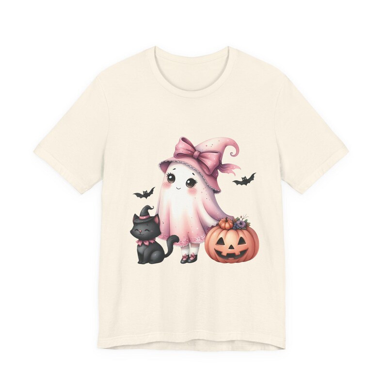 May include: Cream-colored t-shirt featuring a whimsical Halloween design. The graphic includes a ghost wearing a pink witch hat and a black cat with a witch hat, standing next to a carved pumpkin. Bats are flying around the ghost.