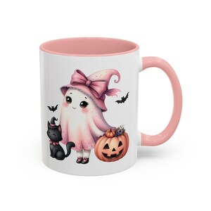 May include: White ceramic mug with a pink interior and handle. The mug features a Halloween-themed illustration of a ghost wearing a pink witch hat and holding a carved pumpkin. A black cat and bats are also depicted.