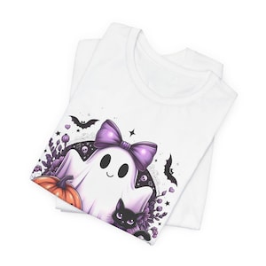 May include: White t-shirt featuring a cartoon ghost with a purple bow, surrounded by a pumpkin, black cat, and bats. The design has a Halloween theme with purple and orange accents.