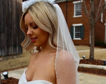 May include: A bride wearing a white strapless wedding dress with a white veil with a beaded edge. The veil is draped over her head and shoulders. She is standing in front of a brick building with a green door.