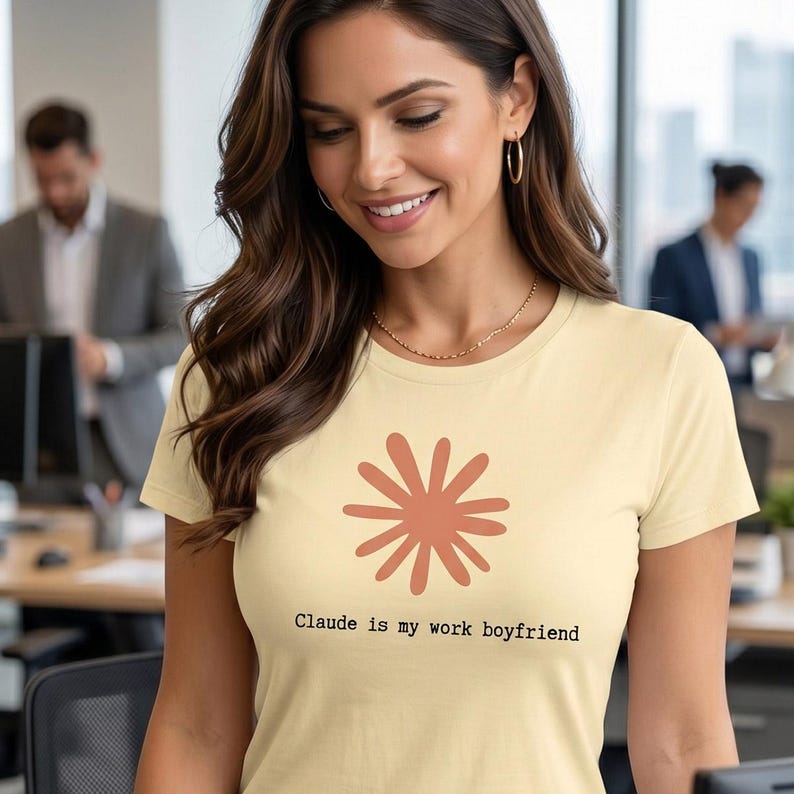 May include: A light yellow t-shirt with a rust-colored floral design and the text "Claude is my work boyfriend." The shirt is made of a soft material and has short sleeves. The person in the image is smiling.