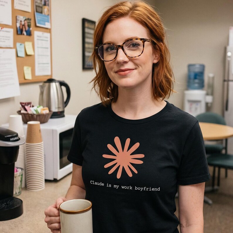 May include: Black t-shirt with a coral-colored floral design and the text "Claude is my work boyfriend." The shirt is worn by a person with red hair and glasses, holding a coffee mug in a kitchen setting.