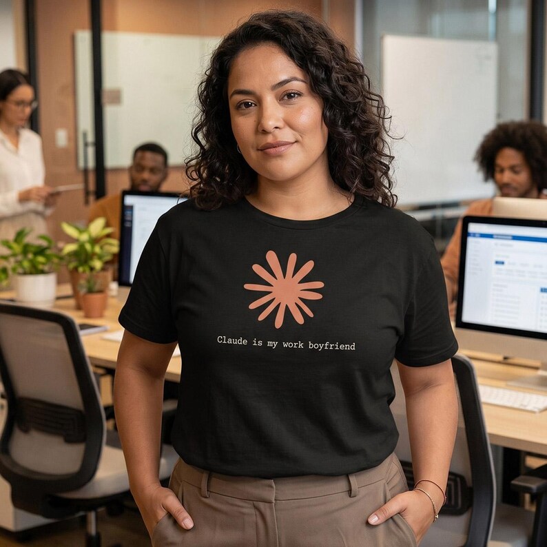 May include: Black t-shirt with a coral-colored flower design and the text "Claude is my work boyfriend." The shirt is a crew neck and is being worn by a person in an office setting.