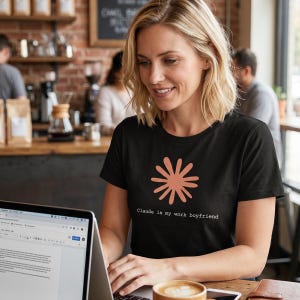 May include: Black t-shirt with a coral-colored floral design and the text "Claude is my work boyfriend." The woman is sitting at a table with a laptop and a cup of coffee in a cafe setting.