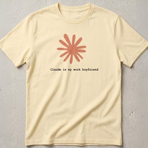 May include: Cream-colored t-shirt featuring a rust-colored floral design and the text "Claude is my work boyfriend." The shirt has a crew neck and short sleeves. The design is centered on the front of the shirt.