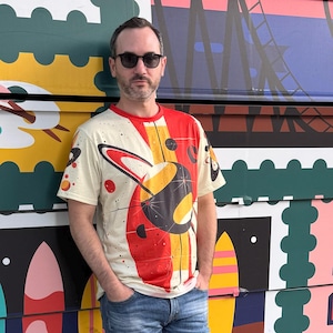 May include: A short-sleeved crew neck t-shirt in cream featuring a large graphic print of a stylised planet with rings in red, yellow, and black. The design includes small starburst accents and geometric shapes across the front, paired with blue denim jeans.