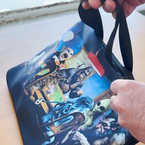 Op de afbeelding: Een zwarte tote bag met rits met een kleurrijke print van een groep personages die kaarten spelen. De personages omvatten een kat, een aap, een zeemeermin, een alien en een schepsel met vacht. De tas heeft twee zwarte banden.