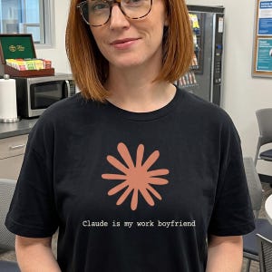 May include: Black t-shirt featuring a coral-colored floral design and the text "Claude is my work boyfriend." The shirt is made of a soft material and has a classic crew neck.