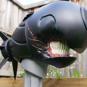 Könnte beinhalten: Ein mattschwarzer, von einer Kreatur inspirierter Helm mit einer abgerundeten, glatten Außenseite und einem markanten Lautsprechergitter an der Vorderseite. Das Design zeigt ein weit geöffnetes Maul mit realistischen weißen Zähnen und rotem Zahnfleisch sowie spitzen Dornen an der Rückseite.