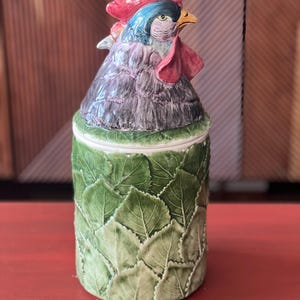May include: A ceramic rooster-shaped jar with a lid. The rooster has a red comb and wattle, blue and green head feathers, and purple body feathers. The jar's base is green and decorated with leaf patterns.