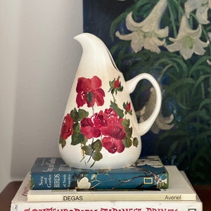 Può includere: Brocca in ceramica bianca con un motivo floreale di rose rosse. La brocca è appoggiata su tre libri, tra cui uno intitolato "Stampe giapponesi contemporanee".