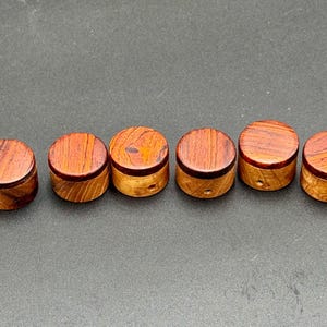 Könnte beinhalten: Sechs kleine, runde Holzkisten mit Deckeln sind in einer Reihe angeordnet. Die Deckel und Oberseiten der Kisten sind rotbraun, während die Seiten hellgelbbraun sind. Jede Kiste hat ein kleines Loch an der Seite.