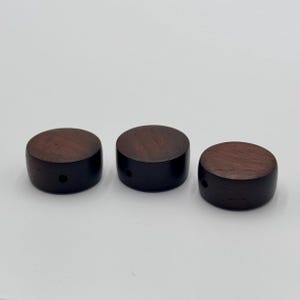 May include: Three dark brown wooden knobs with a circular top and a cylindrical base. Each knob has a small hole drilled through the side. The wood grain is visible on the top surface.