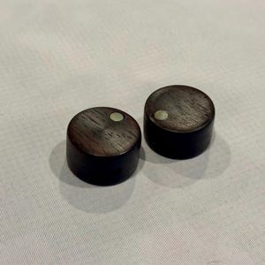 Könnte beinhalten: Zwei dunkelbraune Holzknöpfe mit einem kleinen, runden, hellfarbigen Inlay oben. Die Knöpfe sind zylindrisch und haben eine glatte, polierte Oberfläche. Sie sind wahrscheinlich für Möbel oder Elektronik.